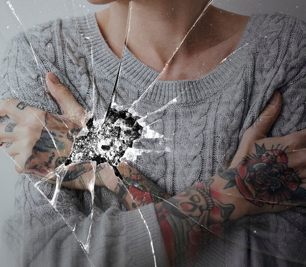 A tattooed woman raises her arms in front of a shattered window.